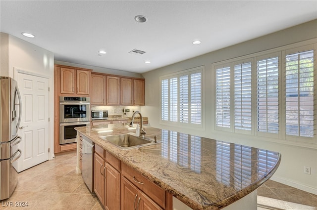 kitchen featuring light tile patterned flooring, a center island with sink, appliances with stainless steel finishes, light stone countertops, and sink