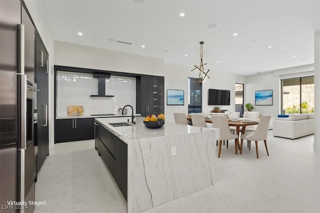 kitchen featuring decorative backsplash, wall chimney exhaust hood, sink, decorative light fixtures, and an island with sink