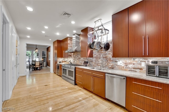 kitchen featuring sink, appliances with stainless steel finishes, hanging light fixtures, wall chimney range hood, and light wood-type flooring