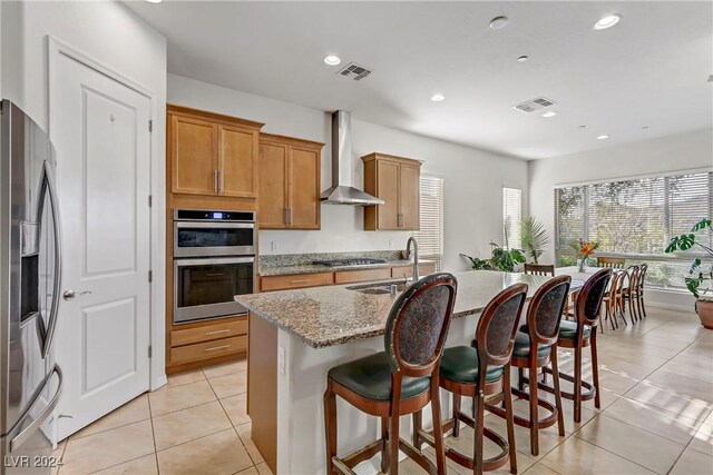 kitchen with wall chimney exhaust hood, sink, a center island with sink, light tile patterned floors, and stainless steel appliances