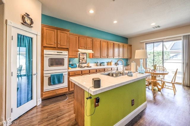 kitchen with tile countertops, white double oven, a center island with sink, sink, and dark hardwood / wood-style flooring