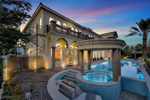 back house at dusk with a swimming pool with hot tub, a balcony, a patio, and exterior kitchen