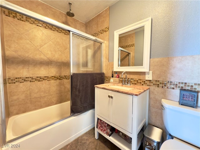 full bathroom with bath / shower combo with glass door, a textured ceiling, toilet, vanity, and tile walls