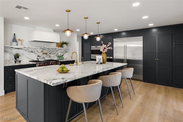 kitchen with appliances with stainless steel finishes, a large island, light stone countertops, custom exhaust hood, and light hardwood / wood-style flooring