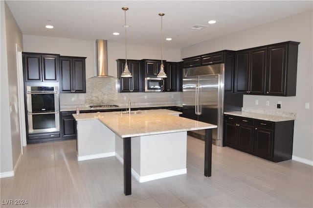 kitchen featuring appliances with stainless steel finishes, wall chimney exhaust hood, a kitchen island with sink, pendant lighting, and a breakfast bar area