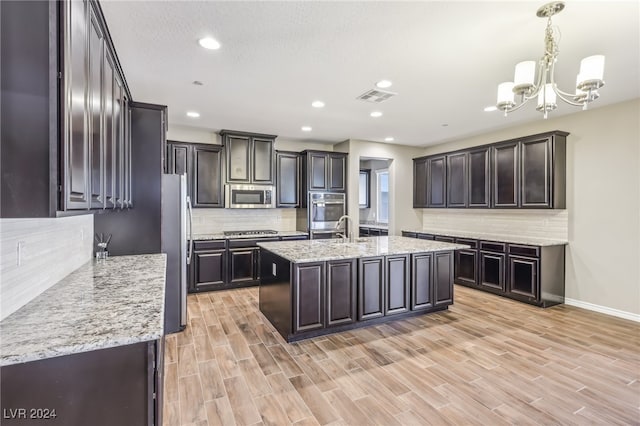 kitchen with dark brown cabinetry, appliances with stainless steel finishes, an island with sink, hanging light fixtures, and light hardwood / wood-style flooring