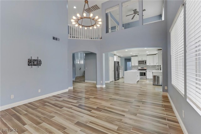 unfurnished living room with ceiling fan with notable chandelier, a high ceiling, and light hardwood / wood-style flooring