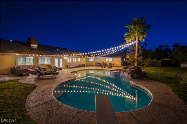 pool at night featuring a lawn, an outdoor living space with a fire pit, a diving board, and a patio area