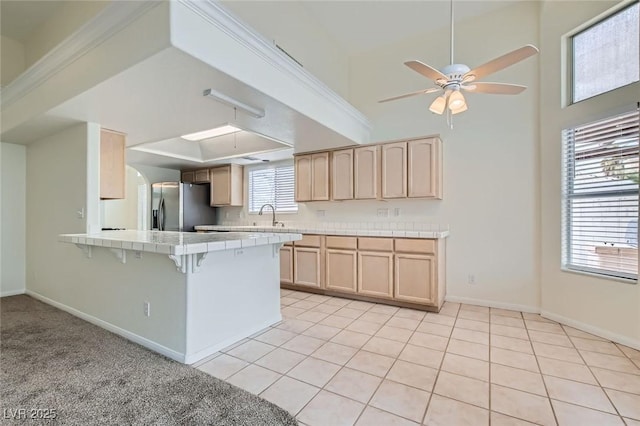 kitchen featuring a peninsula, stainless steel fridge with ice dispenser, light brown cabinetry, tile counters, and a kitchen bar