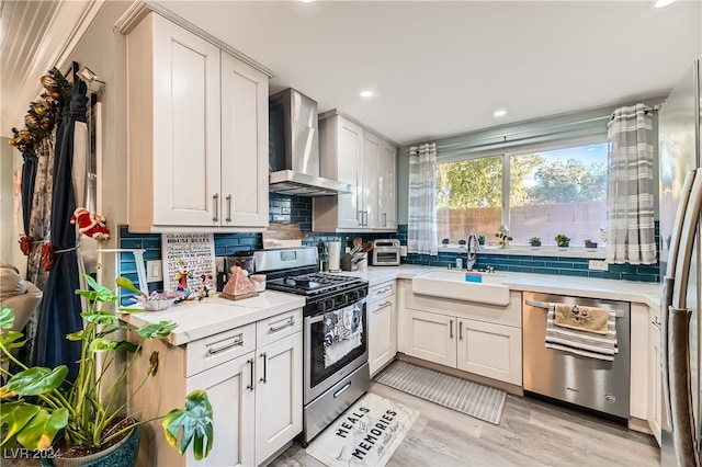 kitchen featuring stainless steel appliances, sink, white cabinets, wall chimney range hood, and light wood-type flooring