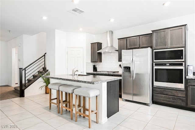 kitchen with sink, an island with sink, wall chimney range hood, a breakfast bar, and appliances with stainless steel finishes