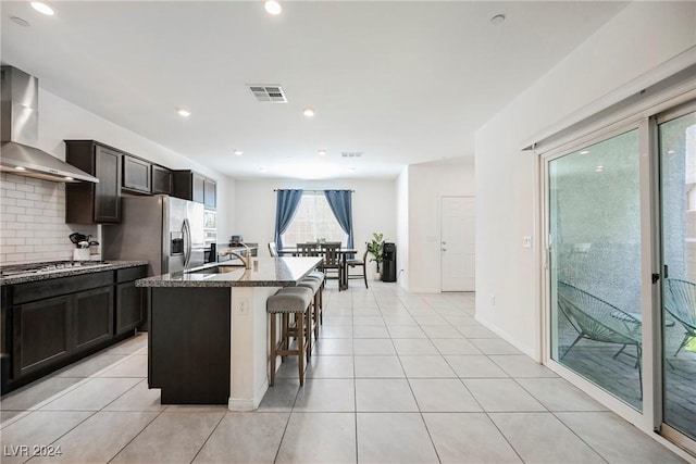 kitchen with light tile patterned flooring, a kitchen bar, a center island with sink, appliances with stainless steel finishes, and wall chimney range hood