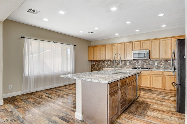 kitchen with light wood-type flooring, appliances with stainless steel finishes, light stone countertops, sink, and a kitchen island with sink