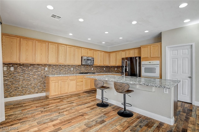 kitchen with light stone counters, a center island with sink, appliances with stainless steel finishes, decorative backsplash, and light hardwood / wood-style flooring