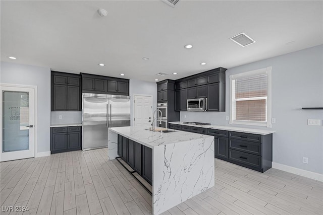 kitchen with light stone counters, stainless steel appliances, sink, a center island with sink, and light hardwood / wood-style floors