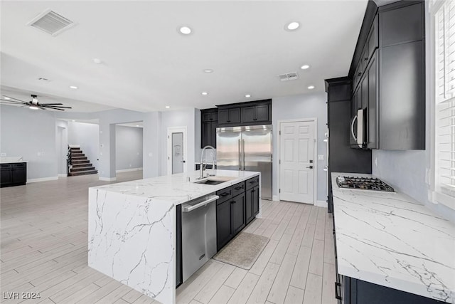 kitchen with sink, ceiling fan, an island with sink, appliances with stainless steel finishes, and light stone counters