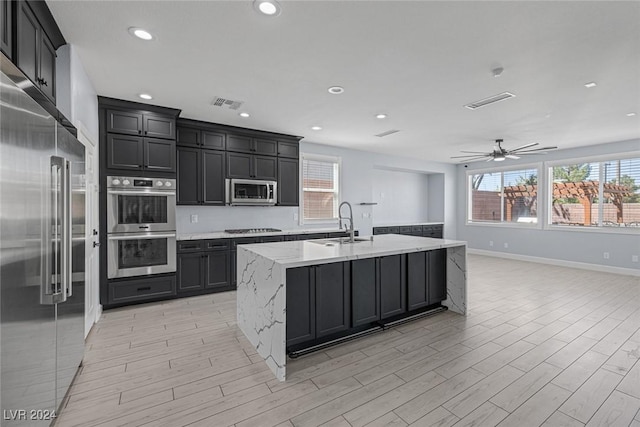 kitchen featuring appliances with stainless steel finishes, light wood-type flooring, light stone counters, ceiling fan, and a center island with sink