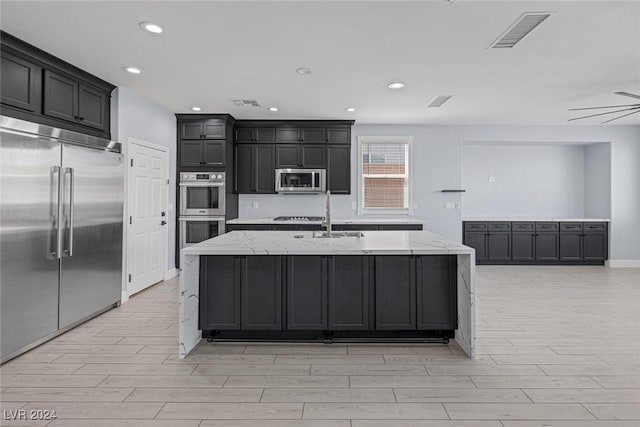 kitchen with light stone countertops, sink, ceiling fan, a kitchen island with sink, and appliances with stainless steel finishes