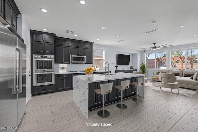 kitchen with appliances with stainless steel finishes, a kitchen breakfast bar, a kitchen island with sink, ceiling fan, and light hardwood / wood-style flooring