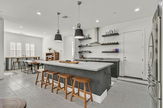 kitchen with appliances with stainless steel finishes, wall chimney exhaust hood, a kitchen island with sink, pendant lighting, and light tile patterned flooring