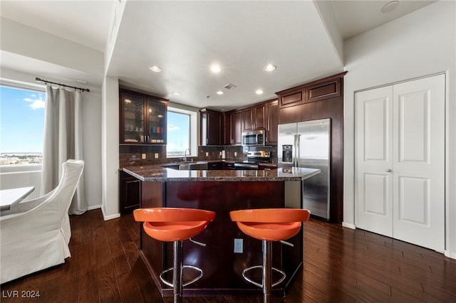 kitchen with appliances with stainless steel finishes, dark hardwood / wood-style flooring, tasteful backsplash, dark stone counters, and a kitchen island