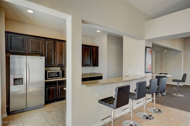 kitchen with light stone countertops, stainless steel appliances, a kitchen bar, dark brown cabinets, and light tile patterned flooring