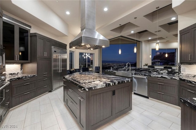 kitchen with dark brown cabinets, appliances with stainless steel finishes, island exhaust hood, dark stone counters, and backsplash