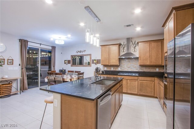 kitchen with stainless steel appliances, wall chimney range hood, sink, pendant lighting, and a center island with sink