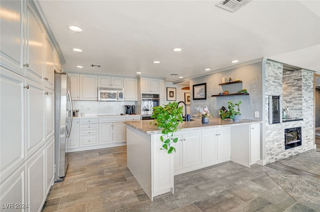 kitchen with light stone countertops, appliances with stainless steel finishes, backsplash, a large fireplace, and white cabinetry