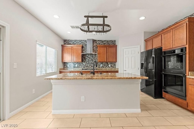 kitchen featuring light stone counters, black refrigerator with ice dispenser, a center island with sink, double oven, and wall chimney range hood