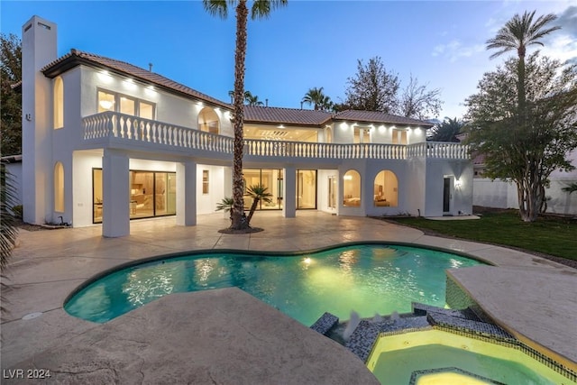 back house at dusk with a balcony, a pool with hot tub, and a patio