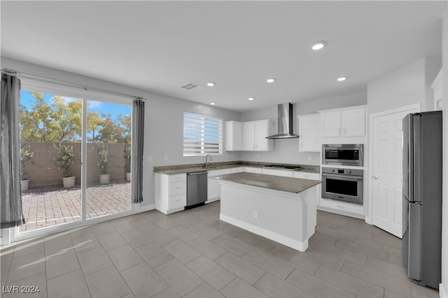 kitchen with wall chimney range hood, light stone countertops, appliances with stainless steel finishes, a kitchen island, and white cabinetry