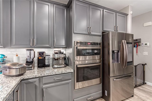 kitchen with appliances with stainless steel finishes, gray cabinetry, decorative backsplash, light stone countertops, and light wood-type flooring