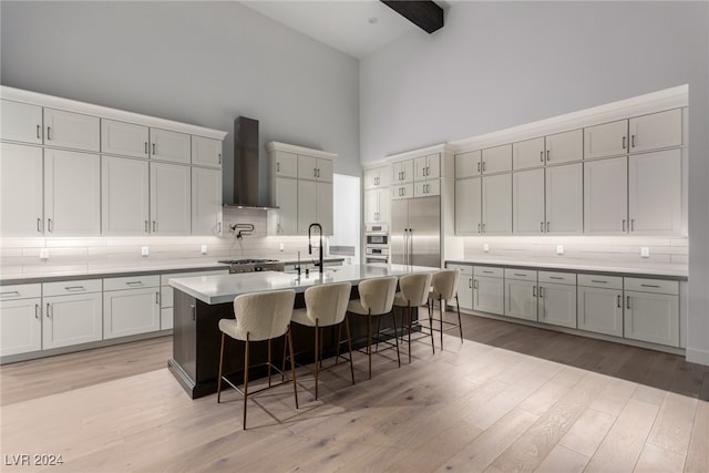 kitchen featuring wall chimney range hood, light hardwood / wood-style flooring, high vaulted ceiling, a breakfast bar, and a center island with sink