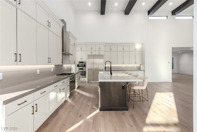 kitchen with wall chimney exhaust hood, light hardwood / wood-style flooring, beamed ceiling, an island with sink, and a breakfast bar area