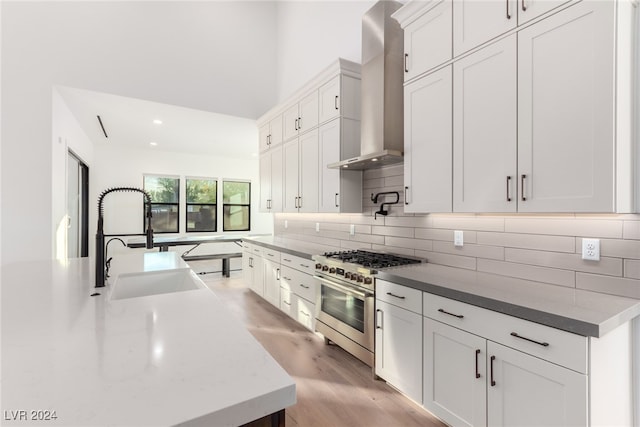 kitchen featuring light wood-type flooring, high end stove, sink, wall chimney range hood, and white cabinetry