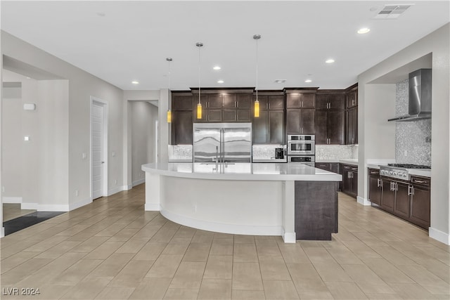 kitchen with appliances with stainless steel finishes, wall chimney exhaust hood, an island with sink, and hanging light fixtures