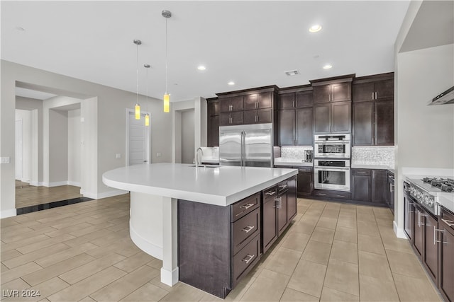 kitchen featuring dark brown cabinetry, hanging light fixtures, stainless steel appliances, tasteful backsplash, and an island with sink