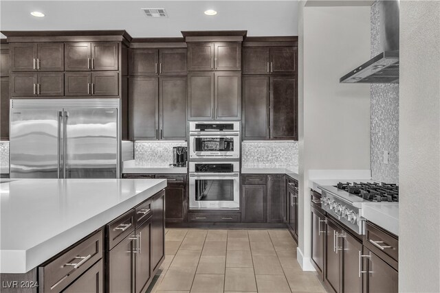 kitchen with backsplash, wall chimney range hood, light tile patterned flooring, dark brown cabinetry, and stainless steel appliances