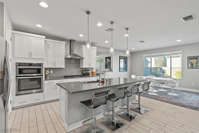 kitchen with white cabinetry, hanging light fixtures, wall chimney range hood, stainless steel appliances, and a kitchen island with sink