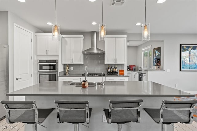 kitchen with white cabinets, wall chimney exhaust hood, stainless steel double oven, and decorative light fixtures