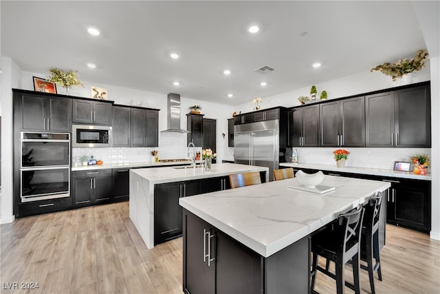 kitchen with decorative backsplash, a kitchen island with sink, wall chimney range hood, built in appliances, and light hardwood / wood-style floors