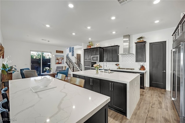 kitchen featuring wall chimney exhaust hood, light wood-type flooring, light stone countertops, an island with sink, and stainless steel appliances