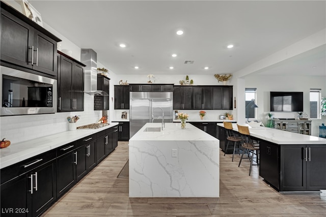 kitchen featuring wall chimney exhaust hood, a kitchen breakfast bar, light hardwood / wood-style flooring, a large island with sink, and built in appliances