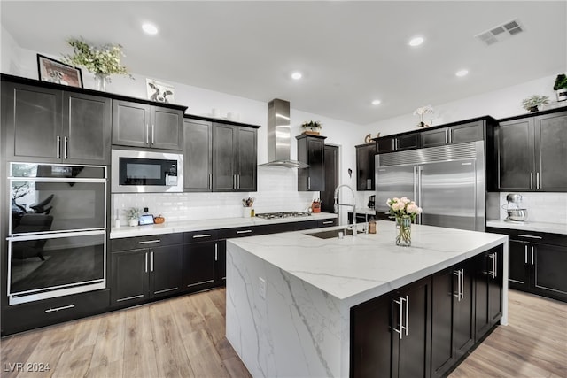 kitchen with sink, built in appliances, wall chimney exhaust hood, an island with sink, and light hardwood / wood-style floors