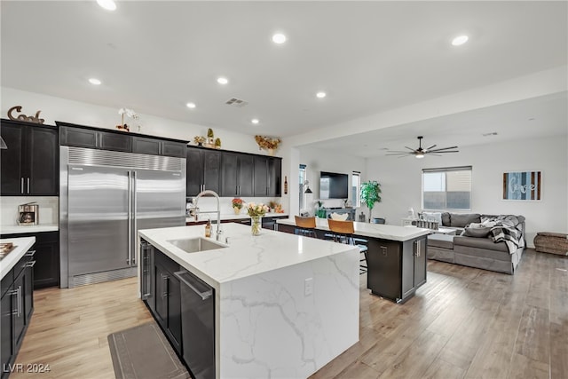 kitchen with light stone counters, stainless steel appliances, a kitchen island with sink, sink, and light hardwood / wood-style floors