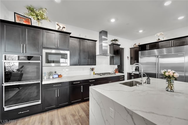 kitchen featuring light stone countertops, sink, wall chimney range hood, built in appliances, and light hardwood / wood-style flooring