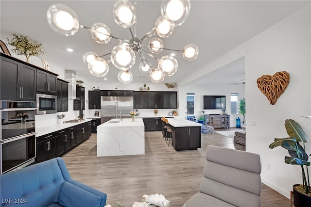 kitchen with ceiling fan with notable chandelier, stainless steel appliances, pendant lighting, light hardwood / wood-style floors, and an island with sink