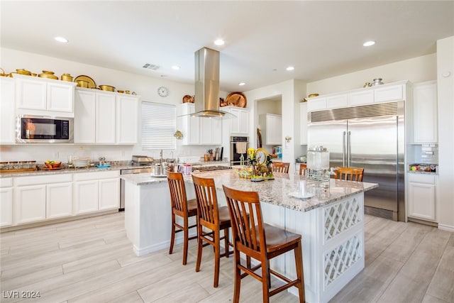 kitchen featuring island exhaust hood, a kitchen bar, light stone counters, a kitchen island with sink, and built in appliances