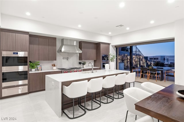 kitchen featuring tasteful backsplash, an island with sink, wall chimney exhaust hood, and stainless steel appliances